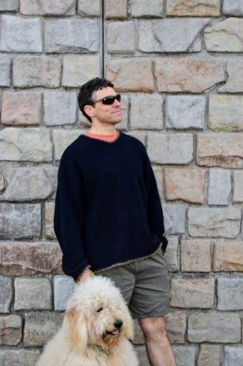 A man stands looking to the right. He is wearing sunglasses and shorts with a long sleeve. There is a fuzzy dog sitting in front of him also looking to the right. There is a brick wall in the background.