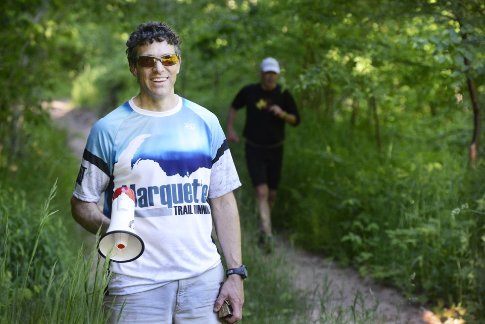 A man in a blue shirt and sunglasses smiling while holding a microphone. They are standing in the woods with trees in the background and another man in black running in the back.