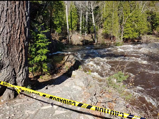 There is water flooding over the rocky trail in the woods. There is a strand of yellow caution tape in front of the water to keep runners from crossing.