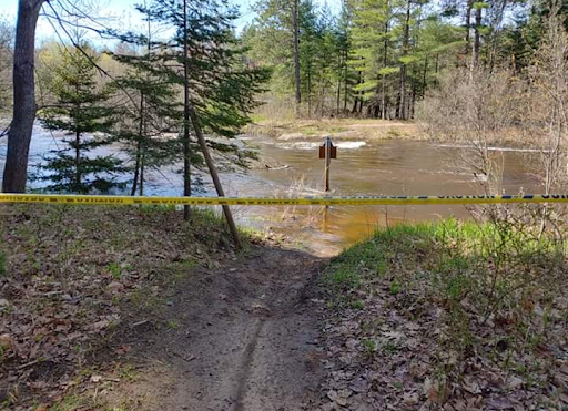 Water is flooding over the trail in the woods covering most of the ground. There are few trees and a strand of caution tape to keep bikers from entering. A map on a post is in the middle of the flood, where trail used to be.