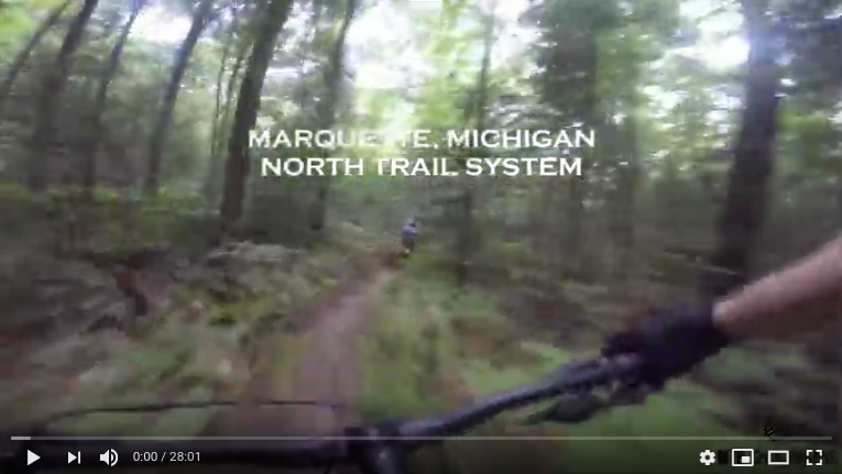 The picture is a screenshot of a YouTube video. It shows a trail in the woods and the handlebars of a bike; from the perspective of the biker. There is text over the image saying 'Marquette, Michigan North Trail System'.